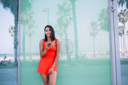 Photo for Cheerful ethnic female in casual clothes messaging on mobile phone and leaning on glass wall while looking at camera and standing near modern building - Royalty Free Image