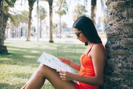 Photo for Side view of serious ethnic female tourist in sunglasses sitting on lawn and planning strategy of trip while leaning on tree and reading paper map - Royalty Free Image