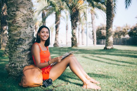 Lächelnde ethnische Touristin mit Rucksack und Fotokamera, die auf einer Landkarte die Richtung überprüft, während sie auf dem grünen Rasen unter Palmen sitzt und in die Kamera blickt