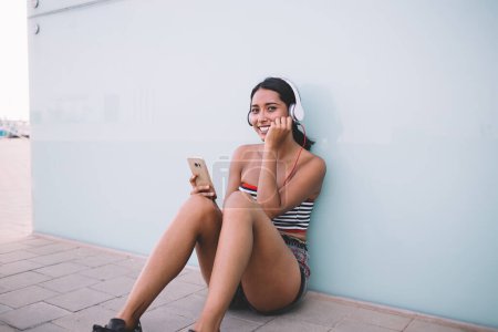 Photo for Smiling ethnic female hipster in summer clothes sitting in headphones on sidewalk and listening to tunes while leaning on glass wall and looking at camera - Royalty Free Image