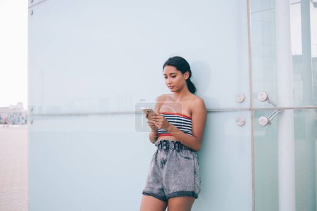 Téléchargez les photos : Jeune brune mince en haut de l'été et smartphone de navigation courte tout en se tenant au mur du bâtiment moderne dans la rue de la ville - en image libre de droit