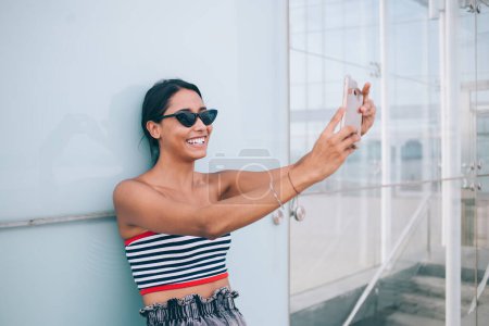Téléchargez les photos : Heureuse brune ethnique dans des lunettes de soleil élégantes et haut en utilisant un téléphone portable et en prenant selfie contre la paroi de verre du bâtiment contemporain - en image libre de droit