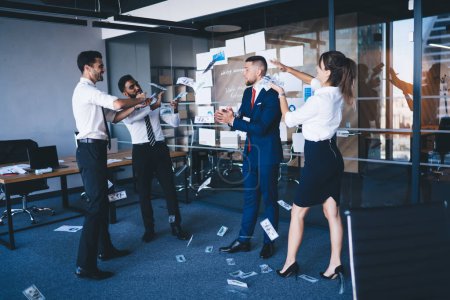 Prosperous male corporate director in formal suit standing in center of room and enjoying monetary rain with employees holding dollars in hand and tossing up, concept of wealthy and abundance
