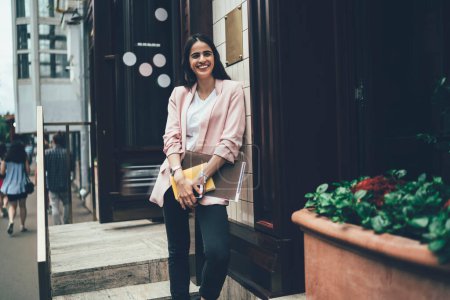 Joyeux hipster fille vêtue de vêtements à la mode tenant matériel d'éducation debout près du bâtiment urbain et souriant, étudiante prospère avec carnet de croquis et bloc-notes riant à la rue de la ville