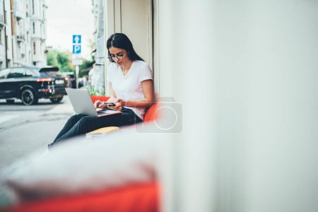 Espagnole femme avec des compétences informatiques lecture reçu un e-mail sur gadget cellulaire moderne, attrayant pigiste féminine dans les lunettes classiques en utilisant un ordinateur portable et des appareils de téléphonie cellulaire pour les réseaux sociaux sur les loisirs