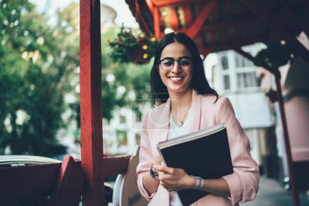Portrait à mi-longueur d'une journaliste féminine joyeuse avec carnet de croquis posant en milieu urbain, femme géorgienne à succès dans des lunettes classiques pour la correction des yeux souriant à la caméra pendant ses loisirs en ville