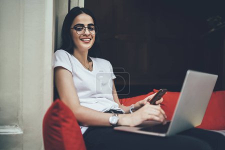 Foto de Retrato de una bonita redactora femenina en anteojos sonriendo a la cámara mientras hace un trabajo remoto en una computadora portátil moderna, mujer hispana feliz con anteojos sosteniendo una computadora digital netbook y posando - Imagen libre de derechos