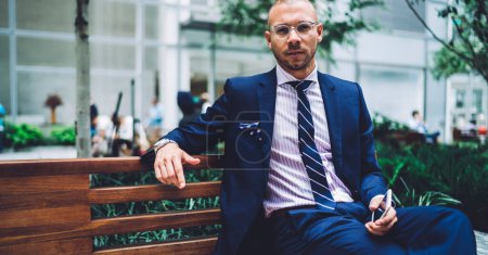 Kaukasischer Professor in luxuriöser eleganter Kleidung und bifokaler Brille, der auf einer städtischen Bank im Stadtzentrum ruht, formell gekleideter erwachsener Student mit Handy in der Hand, der in die Kamera blickt