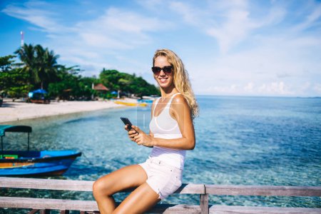 Foto de Sonriente mujer rubia joven bronceada en pantalones cortos blancos y gafas de sol sentados en barandilla de madera a la orilla del mar y tocando en el móvil - Imagen libre de derechos