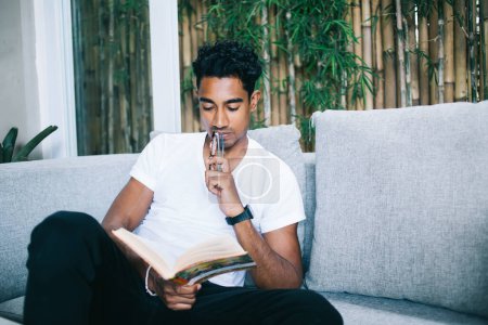 Concentrated young male with holding glasses near mouth thoughtfully reading interesting intellectual book while sitting on comfortable couch at home