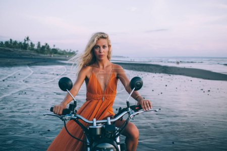 Photo for Attractive young female with long hair in orange dress sitting on motorcycle and looking at camera on seashore at dusk - Royalty Free Image