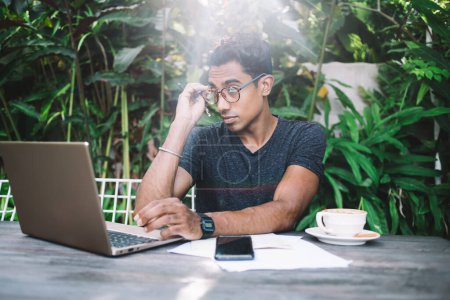 Téléchargez les photos : Homme hispanique concentré dans les lunettes et t-shirt casual navigation ordinateur portable tout en étant assis à la table avec des documents et tasse de café au café de la rue - en image libre de droit