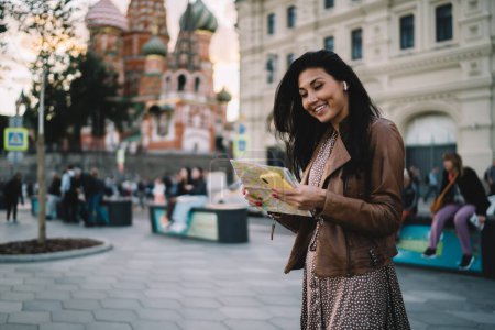 Foto de Feliz turista asiático en auriculares bluetooth que exploran la ciudad de Moscú utilizando el dispositivo de teléfono celular y el mapa de papel para rastrear la ubicación de GPS durante el recorrido turístico, chica alegre en auriculares disfrutando de viaje en solitario - Imagen libre de derechos