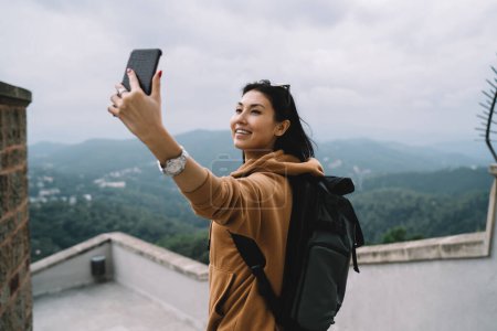 Foto de Sonriente mujer asiática turista en ropa casual y mochila haciendo foto en la cámara del teléfono móvil durante las vacaciones, mujer alegre posando para selfie explorar destinos en viaje chare content online - Imagen libre de derechos