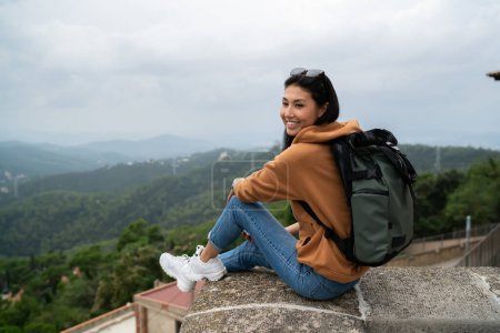 Fröhliche asiatische Reiseleiterin in Freizeitkleidung und trendigem Rucksack, die am Ziel ruht, Sehenswürdigkeiten auf der Reise entdeckt, lächelnde Touristin, die sich auf Sightseeing-Tour im Urlaub erholt