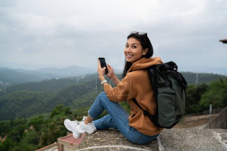 Rear view of asian female travel blogger holding mobile phone sending messages during rest on trip on historical place, smiling woman millennial tourist enjoying visiting location use application