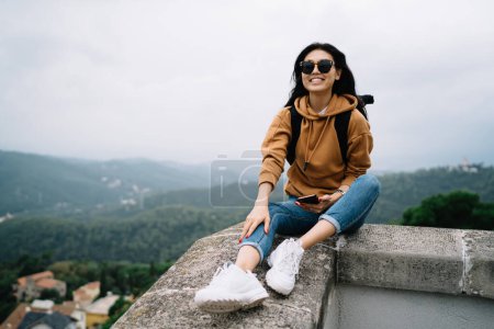 Fröhliche asiatische Reisende mit trendiger Sonnenbrille genießen Reise und Fernweh im Urlaub, lächelnde Frau sitzt am Ziel und ruht während einer Sightseeingtour Ziele auf der Reise entdecken