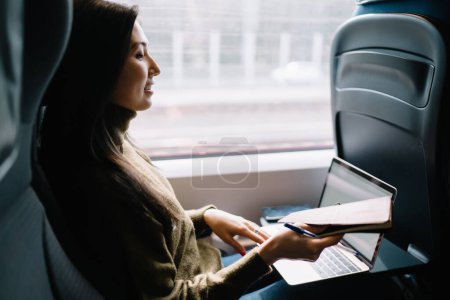 Photo for Positive asian female journalist working remotely during journey thinking about publication for web page holding notebook in train, positive woman using laptop computer for research in modern train - Royalty Free Image