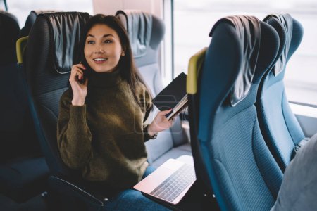 Foto de Alegre mujer de negocios asiática hablando por teléfono móvil mientras viaja en tren con tecnologías modernas y conexión inalámbrica a Internet, sonriente mujer próspera haciendo llamada de teléfono inteligente en viaje - Imagen libre de derechos