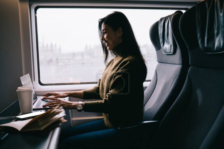 Photo for Cheerful asian woman blogger typing on laptop computer working remotely during trip in train satisfied with connection,smiling female digital nomad keyboarding and watching video in wagon on trip - Royalty Free Image