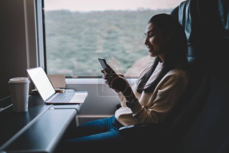 Foto de Mujer asiática sonriente leyendo mensaje de ingreso en teléfono móvil trabajando a través de tecnología en vagón de tren, bloguera positiva comparte contenido de medios sobre viaje durante transporte ferroviario en celular - Imagen libre de derechos