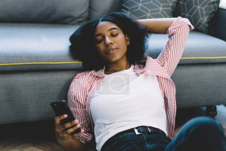 Foto de Mujer afroamericana usando aplicación de teléfono celular para hacer chat en línea durante el fin de semana de ocio en el apartamento en casa, hermosa bloguera escribiendo texto de contenido para su publicación de descarga - Imagen libre de derechos