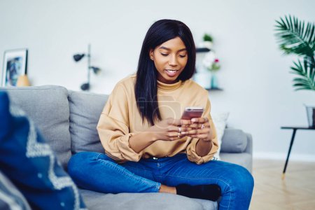 Foto de Mujer de piel oscura disfrutando de la mensajería en línea en la tecnología moderna de teléfonos celulares durante el ocio de fin de semana en el apartamento en casa con un interior acogedor, blogger afroamericano 20s networking social wireless - Imagen libre de derechos