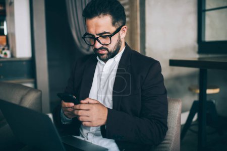 Pensativo empresario masculino caucásico en gafas para la protección de la visión enviar un mensaje en el teléfono móvil, mientras que el trabajo remoto, confiado notificación de comprobación ejecutiva masculina sobre la transacción con el ordenador portátil