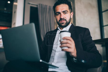 Serious caucasian bearded male entrepreneur having coffee break checking mails on laptop computer, confident businessman in formal wear looking at camera holding cup during remote job in cafe