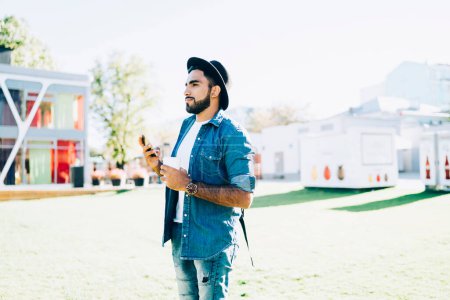 Téléchargez les photos : Vue latérale d'un homme ethnique aux cheveux noirs barbus réfléchi en chapeau et en costume de denim debout dans un parc lumineux avec une tasse de café et regardant ailleurs - en image libre de droit