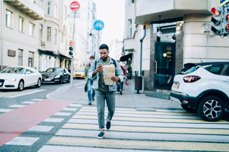 Concentrated bearded ethnic man dressed in casual clothes navigating with paper map and mobile phone going through crosswalk in city at daytime