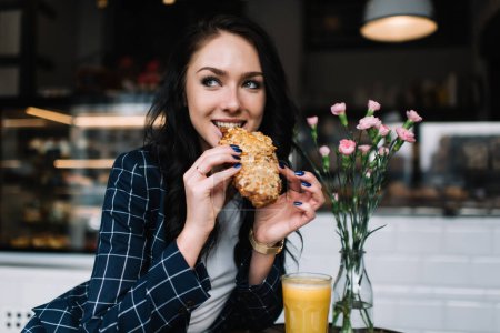 Téléchargez les photos : Joyeux jeune femme souriant et regardant loin tout en étant assis à la table avec un verre de jus de fruits frais et en mangeant un pain dans un café moderne - en image libre de droit