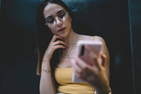 Téléchargez les photos : De dessous de la culture jeune femme portant une tenue décontractée et des lunettes à l'aide d'un téléphone mobile pour prendre selfie à l'extérieur près du mur noir - en image libre de droit