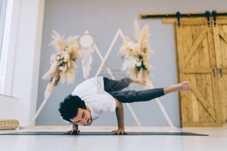 Full length of man in comfortable sportswear doing Eka Pada Koundinyasana asana while stretching body and improving balance during yoga class