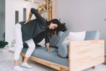 Photo for Side view of woman in home clothes standing bent over and leaning on sofa with hand and experiencing discomfort in back when rising from floor in living room at home - Royalty Free Image