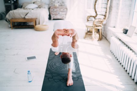 Young male yoga instructor demonstrating yoga Crescent Lounge Halfway Fold pose while training on rubber mat next to window in living room