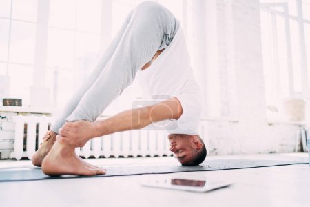Téléchargez les photos : Corps entier de gourou de yoga concentré en vêtements de sport de couleur claire faisant du yoga Prasarita Padottanasana pose tout en se tenant debout sur un tapis en caoutchouc près de la fenêtre en classe lumineuse - en image libre de droit