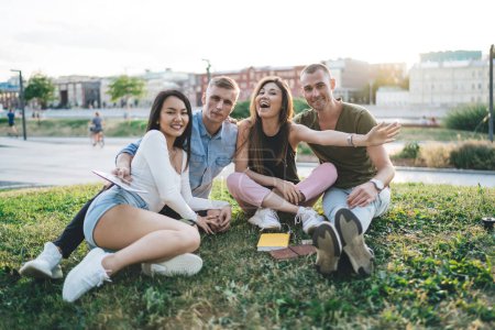 Porträt fröhlicher Studentinnen und Studenten in lässiger Kleidung, die mit Freundschaftsbeziehungen zufrieden sind, fröhliche Hipster, die im Gras sitzen, im Hintergrund die Schule und in die Kamera lächeln