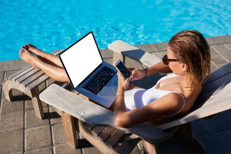 From above of young woman in sunglasses messaging on cellphone and using laptop with blank screen while spending time near swimming pool