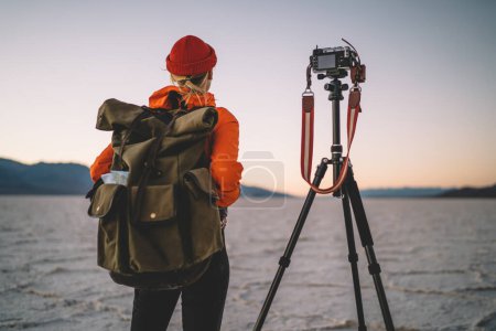 Blick von unten auf einen unkenntlichen Touristen mit Rucksack, der im trockenen Tal steht, während er den Sonnenuntergang mit der Fotokamera aufzeichnet und durch die USA reist