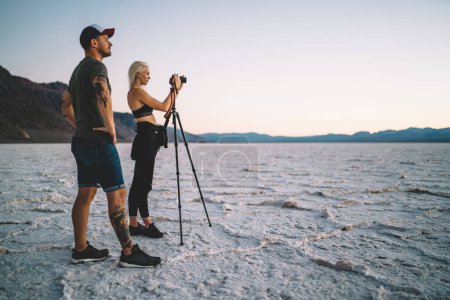 Side view of couple of young photographers taking pictures of Badwater Basin near mountains while exploring Death Valley during road trip across USA