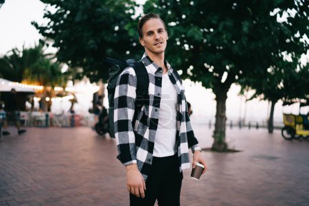 Photo for Half length portrait of positive caucasian male traveler with backpack standing on urban setting city square, young 20s hipster guy looking at camera explore destinations during trip visiting town - Royalty Free Image