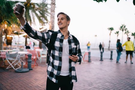 Photo for Smiling caucasian hipster guy in casual trendy wear and with backpack gesture raising hand with coffee to go cup on street, positive male male traveler enjoying exploring city on free time vacations - Royalty Free Image