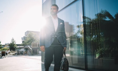 Pensive businessman with trendy briefcase walking at downtown urbanity and thoughtful looking away, middle aged male entrepreneur dressed in luxury outfit feeling pondering on way to office
