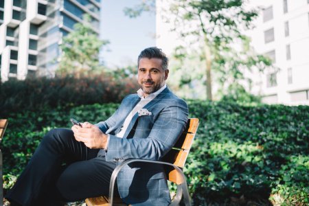 Portrait of mature businessman in elegant apparel looking at camera while resting at urban bench in metropolitan downtown, formally dressed male proud CEO with smartphone gadget in hands posing
