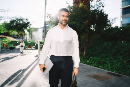 Photo for Successful businessman in formal white shirt walking at sunny urban street and smiling enjoying work break in financial district, confident middle aged male boss with laptop technology and briefcase - Royalty Free Image