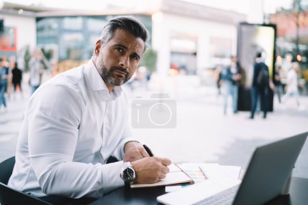 Portrait of mature entrepreneur in formal white shirt looking at camera during time for business planning, middle aged male employer working remotely in sidewalk cafe posing at table with netbook