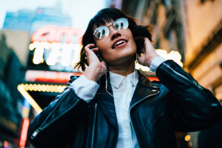 Entspanntes Hipstermädchen, das sich bei guter Musik beim Stadtbummel durch Manhattan erholt, trendig gekleideter Millennial-Teenager in Brille bewundert ruhige Songs, die in elektronischen Stereo-Kopfhörern angehört werden