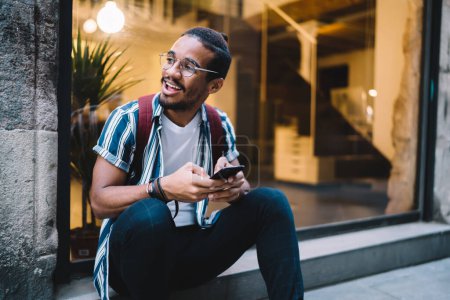 Foto de Positivo chico hipster de piel oscura feliz en gafas mirando hacia otro lado utilizando el teléfono inteligente para comunicarse en tiempo libre en la ciudad, alegre hombre afroamericano sosteniendo el teléfono móvil para navegar en el ocio - Imagen libre de derechos