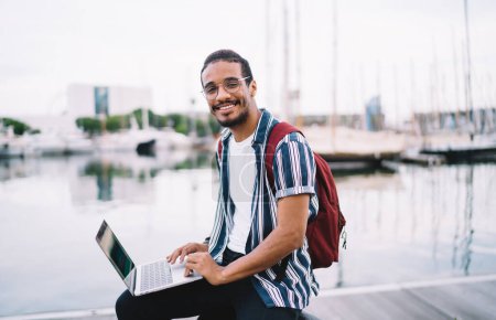 Fröhliche dunkelhäutige männliche Blogger teilen Inhalte von einer Reise mit Laptop-Computer mit 4G-Internetverbindung zufrieden, lächeln fröhlich afrikanisch-amerikanischen Hipster-Kerl schreiben und kommunizieren online